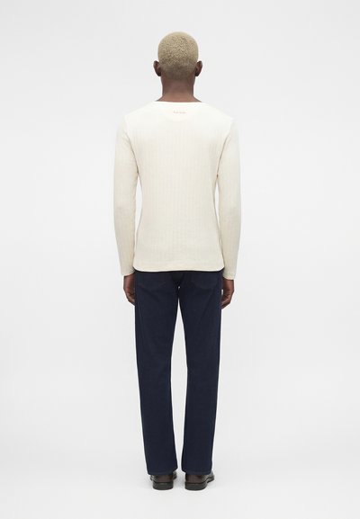 Άτομο με κοντά ξανθά μαλλιά που φοράει εφαρμοστή κρεμ μακρυμάνικη μπλούζα και σκούρα μπλε τζιν, όρθιο με πλάτη προς τα εμπρός σε λευκό φόντο.