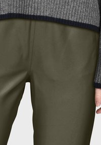 Pantalons ajustés vert olive avec une texture lisse, présentés à côté d'un pull rayé gris et marine, partiellement visible.