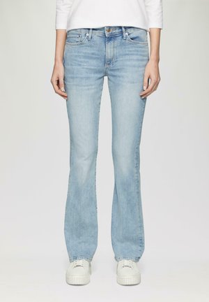 Jean évasé bleu clair en denim, avec un design à cinq poches standard, un léger effet délavé et une taille mi-haute. Porté avec des baskets blanches.