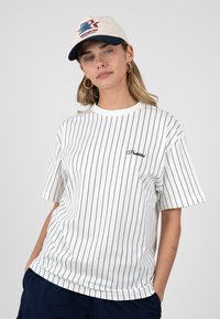 Jeune femme portant un t-shirt blanc à fines rayures avec un logo, un short bleu marine, une casquette beige et bleu marine, et des boucles d’oreilles créoles, debout les mains dans les poches.