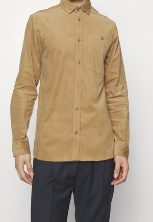 Homme portant une chemise boutonnée en velours côtelé marron clair à manches longues et un pantalon noir, debout devant un fond uni.