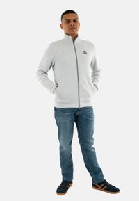 Grijze ritsvest met hoge kraag, geribde manchetten, voorkuipen, gecombineerd met blauwe jeans en zwarte sneakers met bruine accenten.