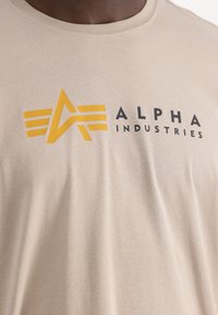 Camiseta beige de algodón con cuello redondo. Presenta un logo amarillo con tres líneas y el texto "ALPHA INDUSTRIES" en negro.