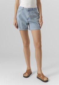 Pantaloni shorts in denim azzurro chiaro con una vestibilità comoda, orli arrotolati, due tasche frontali e chiusura con bottone. Abbinati a sandali scivolati marroni.