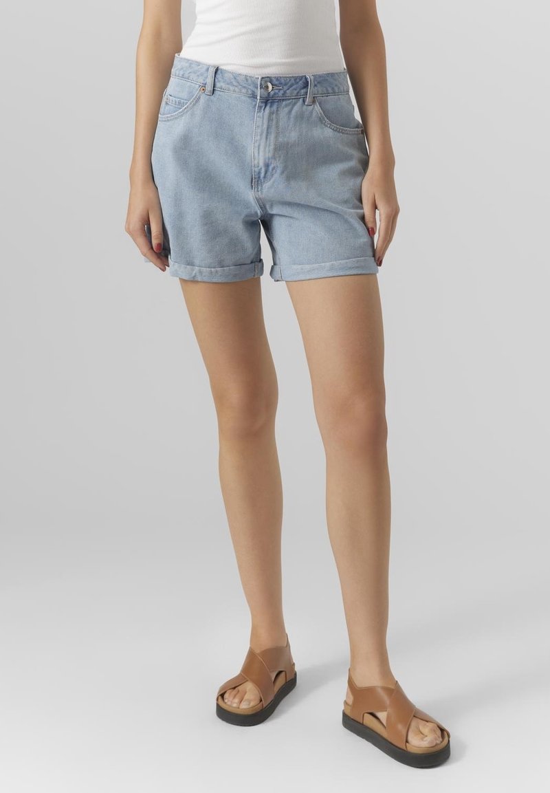 Pantaloni shorts in denim azzurro chiaro con una vestibilità comoda, orli arrotolati, due tasche frontali e chiusura con bottone. Abbinati a sandali scivolati marroni.