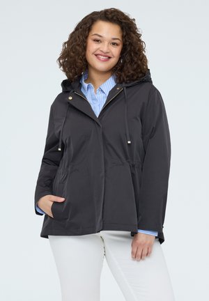 Mujer sonriente con una chaqueta negra con capucha sobre una camisa azul a rayas y pantalones blancos, con una mano en el bolsillo de la chaqueta.