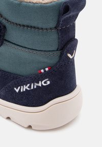 Calzado de ante azul oscuro con parte superior de tejido verde, que presenta el logotipo "VIKING" en blanco, acentuado por una etiqueta roja y blanca. Diseño de suela redondeada.