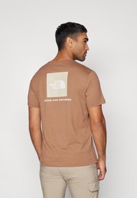 Camiseta de algodón marrón con un diseño de logo cuadrado en beige claro en la parte posterior, con el texto "NUNCA DEJES DE EXPLORAR" debajo.