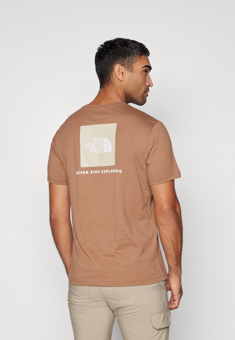 Camiseta de algodón marrón con un diseño de logo cuadrado en beige claro en la parte posterior, con el texto "NUNCA DEJES DE EXPLORAR" debajo.