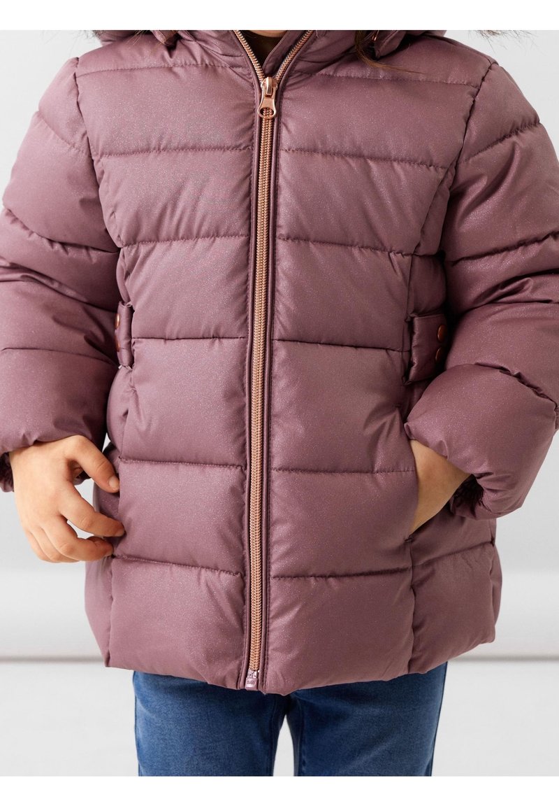 Enfant portant une doudoune mauve avec fermeture éclair devant, mains partiellement dans les poches, associée à un jean bleu sur un fond uni.