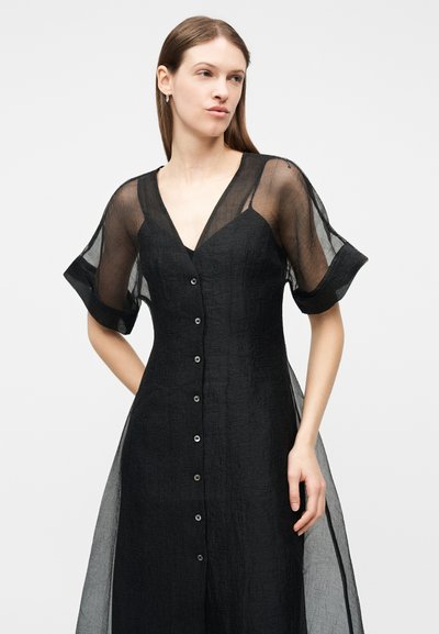 Γυναίκα με μαύρο φόρεμα με κουμπιά, διάφανα κοντά μανίκια και βαθύ V-λαιμόκοψη, στέκεται με το ένα χέρι στη μέση σε απλό φόντο.