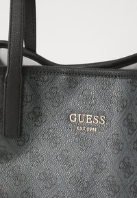 Crna torba od teksturiranog materijala s sjajnim zlatnim "GUESS" logotipom i detaljem "EST 1981". Ima dvije čvrste ručke.
