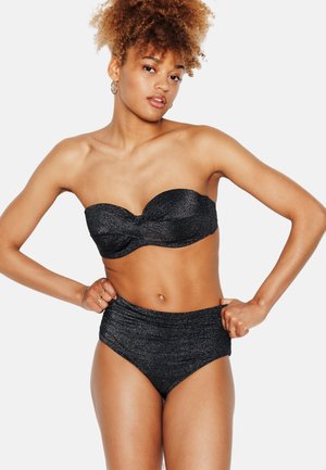 Donna con capelli ricci che indossa un top bikini nero scintillante senza spalline e slip a vita alta, posa con le mani sui fianchi su sfondo bianco.