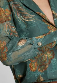 Blusa verde con estampado, hecha de tela transparente, que presenta bordados florales en dorado y naranja, con un detalle de botón en el puño.