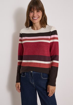 Femme souriante portant un pull à manches longues rayé dans des tons rouge, rose, blanc et marron avec un jean bleu foncé, debout contre un mur uni.