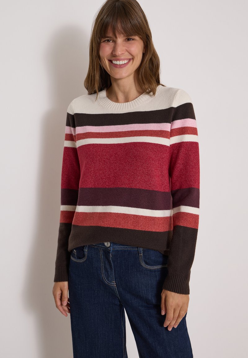 Femme souriante portant un pull à manches longues rayé dans des tons rouge, rose, blanc et marron avec un jean bleu foncé, debout contre un mur uni.
