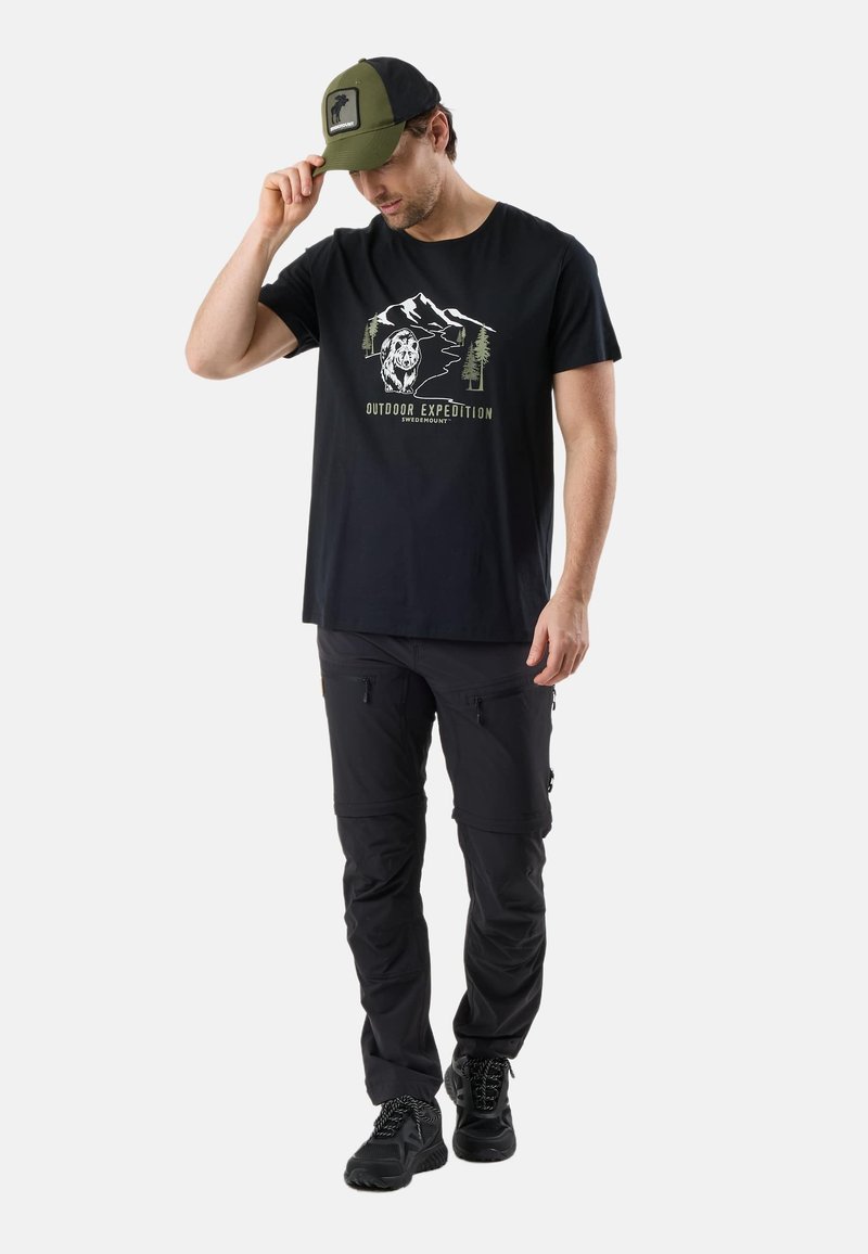 Homme portant un t-shirt noir « Outdoor Expedition » avec un graphisme de montagne et d'ours, un pantalon cargo noir, des chaussures noires et une casquette verte et noire, ajustant son chapeau.