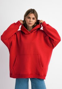 Rode oversized hoodie met een voorzak, dikke manchetten en een grote kap. Smooth textuur en een relaxte pasvorm, gecombineerd met een spijkerbroek.