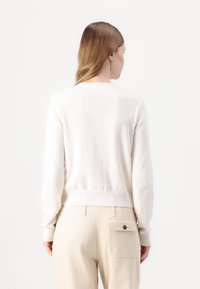 Lys beige strikket genser med rund hals, ribbestrikket kant og lange ermer, brukt over kremfargede bukser med baklomme.