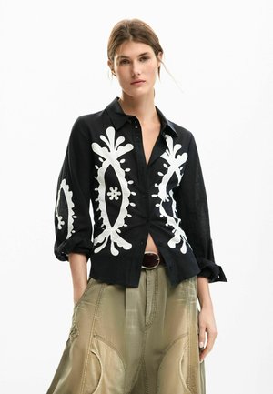 Zwart overhemd met knopen, versierd met witte bloemen- en paisleyborduursels. Lange mouwen, gestructureerde pasvorm, gecombineerd met licht kaki, hooggetailleerde broek.