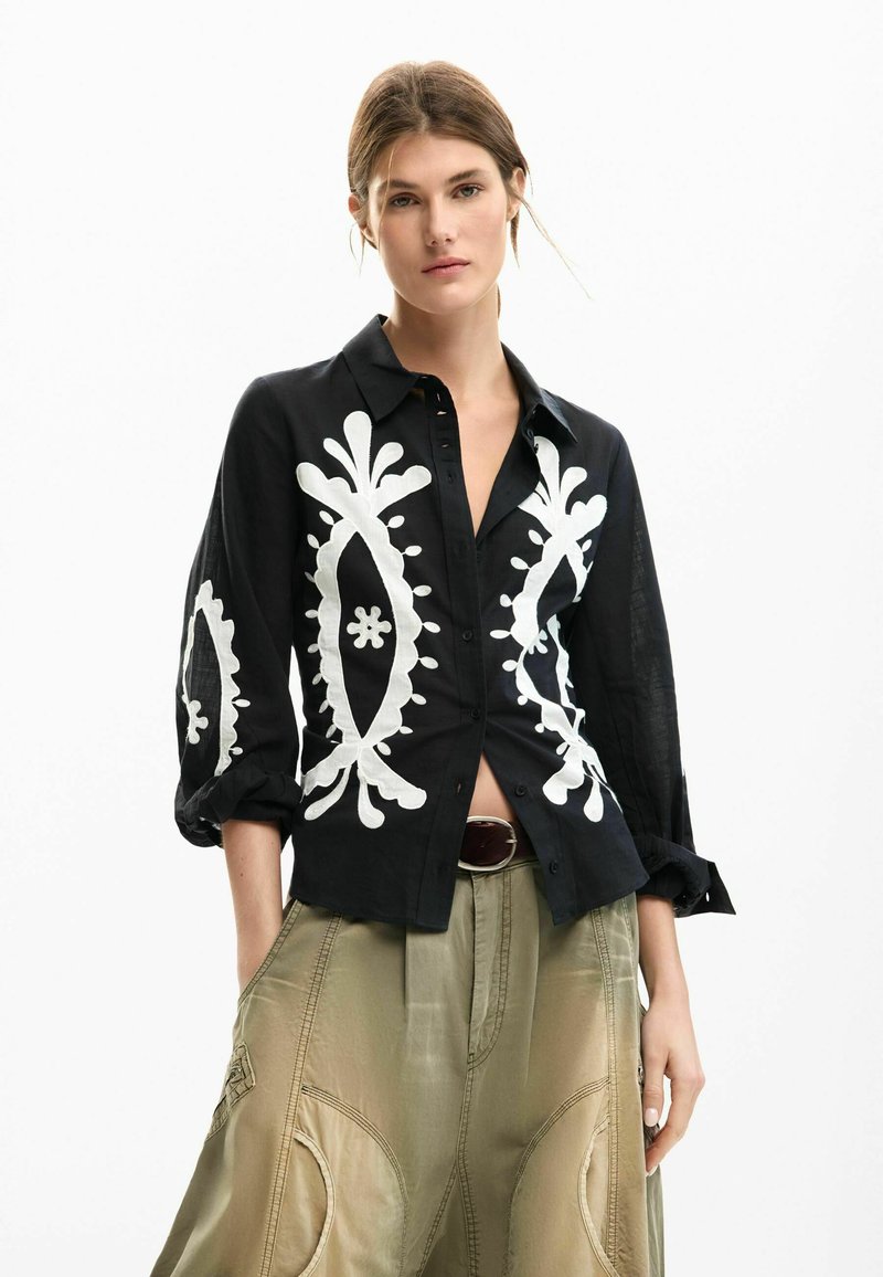 Schwarzes Hemd mit Knopfleiste, verziert mit weißer Blumen- und Paisley-Stickerei. Lange Ärmel, strukturierte Passform, kombiniert mit hellen, hoch taillierten Khakihosen.