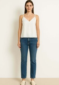 Donna in piedi contro un muro neutro, che indossa un top senza maniche bianco, jeans blu e mocassini color crema.