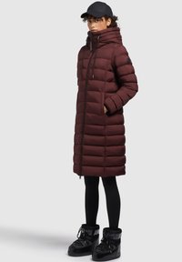 Manteau rembourré marron avec un col haut, fermeture éclair et poches latérales. Associé à des leggings noirs et des bottes noires isolées.