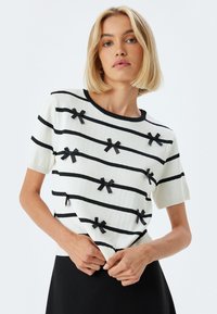 Suéter blanco de punto con rayas horizontales negras y pequeños acentos de lazo negro. Mangas cortas y cuello y dobladillo acanalados.