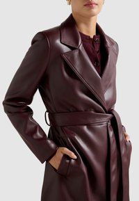 Βουργουνδί δερματίνη τρανς παλτό με ζώνη στη μέση, φαρδιούς γιακάδες, μακριά μανίκια και πλαϊνές τσέπες. Διαθέτει λείο, γυαλιστερό ύφασμα.