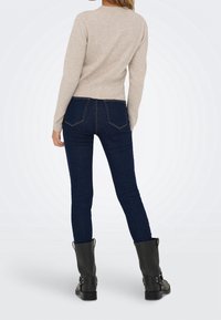 Femme portant un pull beige à manches longues, un jean skinny bleu foncé et des bottes noires mi-mollet, debout, dos à la caméra sur fond blanc.