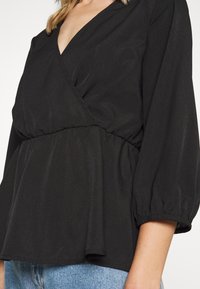 Blusa nera avvolgente con scollatura a V profonda, vita arricciata e maniche voluminose. Texture liscia e tessuto morbido, con orlo fluttuante.