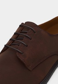 Καφέ δερμάτινο παπούτσι με κλειστή κορδόνια και λεπτά στρογγυλά κορδόνια σε σκούρο καφέ χρώμα, με λεπτομερή ραφή κοντά στα οπές για κορδόνια.