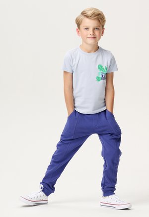 Lichtblauwe T-shirt met korte mouwen en groene bladerenprint, gecombineerd met paarse baggy broek en witte sneakers met rode accenten.