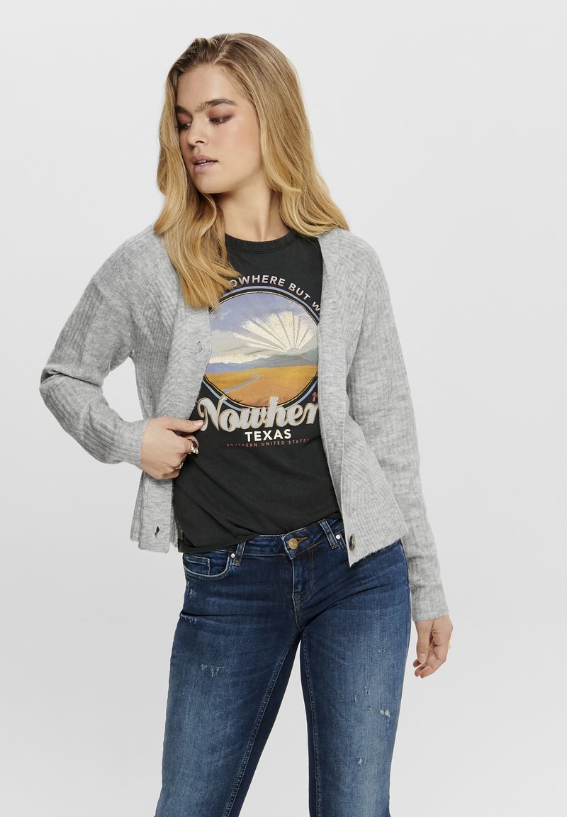 Grau gerippter Cardigan mit lockerer Passform, ausgestattet mit Knöpfen und langen Ärmeln, über einem schwarzen Grafik-T-Shirt getragen und kombiniert mit dunkelbblauen Jeans.