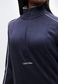 Μπλε ναυτικό τοπ με φερμουάρ, με λευκή λεπτομέρεια στους ώμους και τα μανίκια, λογότυπο Calvin Klein κάτω από τον γιακά με φερμουάρ, φορεμένο από ένα άτομο.