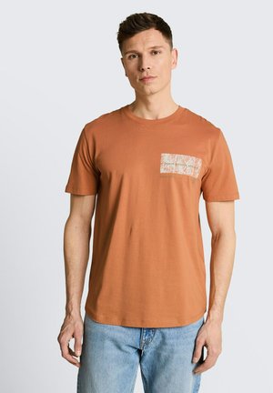 Man draagt een roestkleurig T-shirt met een patroon op de borstzak en lichtblauwe spijkerbroek, staand tegen een effen lichte achtergrond.