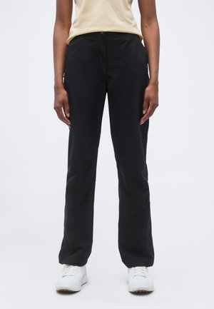 Henkilö, jolla on suorat mustat housut, beige lyhythihainen paita ja valkoiset tennarit, seisoo yksivärisen taustan edessä.