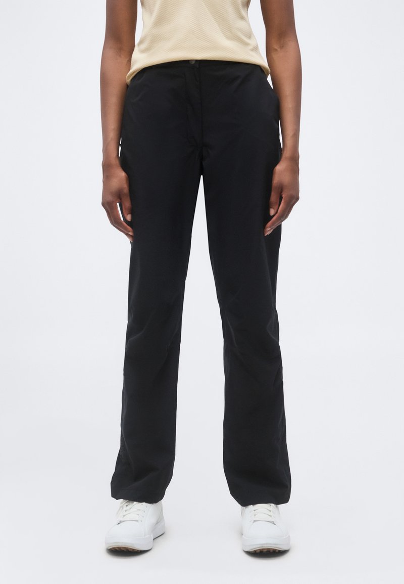 Άτομο που φοράει μαύρο παντελόνι με ίσια γραμμή, μπεζ κοντομάνικο τοπ και άσπρα αθλητικά παπούτσια, στέκεται μπροστά σε ένα μονόχρωμο φόντο.