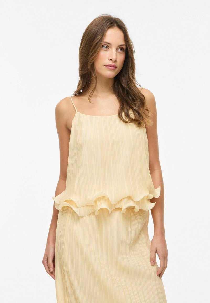 Jeune femme aux cheveux ondulés lâchés, portant une robe sans manches plissée de couleur jaune pâle avec un détail volanté à la taille, debout devant un fond blanc uni.