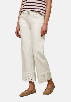 Kobieta ubrana w białe, szerokie spodnie 3/4 z koronką typu ażurowego na dole, paskowany top bez rękawów oraz brązowe zamszowe płaskie buty z klamrą.