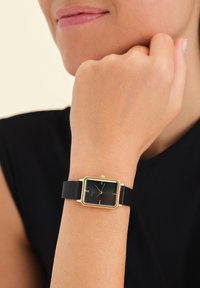 Reloj rectangular negro con un marco de metal dorado y una correa de cuero negro texturizado, que presenta marcadores de hora simples y una corona.
