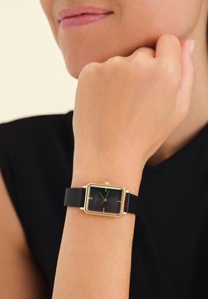 Montre rectangulaire noire avec un cadran en métal doré et un bracelet en cuir noir texturé, présentant de simples marqueurs d'heure et une couronne.