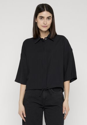 Jeune femme aux cheveux raides et foncés porte une chemise noire ample boutonnée à manches mi-longues et un pantalon noir, debout devant un fond uni.