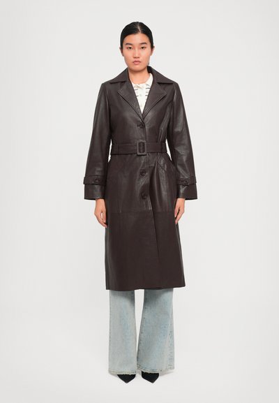 Donna in piedi che indossa un trench coat in pelle marrone scuro con cintura sopra dei jeans a gamba larga blu chiaro e scarpe a punta nere.