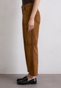 Bruine suede broek met een rechte pasvorm, voorzien van voorzakken, een knoopsluiting en zichtbare stikdetails. Gecombineerd met zwarte instapschoenen.