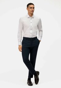 Camicia bianca a maniche lunghe con bottoni neri, abbinata a pantaloni blu navy e scarpe di pelle nere, con una vestibilità sartoriale.
