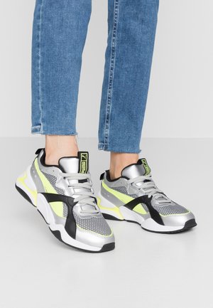 Ben i blå jeans iført sølv, sort og neon-gule Puma-sneakers med snørebånd og mærkede tunge tags på en grå gulv.