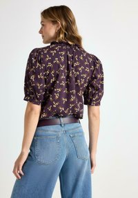 Kortærmet bluse med en mørk lilla bund, prydet med et blomsterdesign i gult og hvidt, flæse ved halsen og puffede ærmer. Bæres med lyseblå viddejeans.