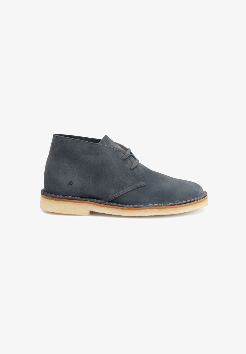 Scarpe chukka in suede grigio scuro con punta arrotondata, due lacci e suola in gomma beige a contrasto. Presentano un delicato dettaglio di cucitura.