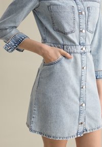 Lichtblauw denim jurk met knoopsluiting, twee borstzakken en een getailleerde taille. Heeft opgerolde mouwen en zichtbare stiksels.